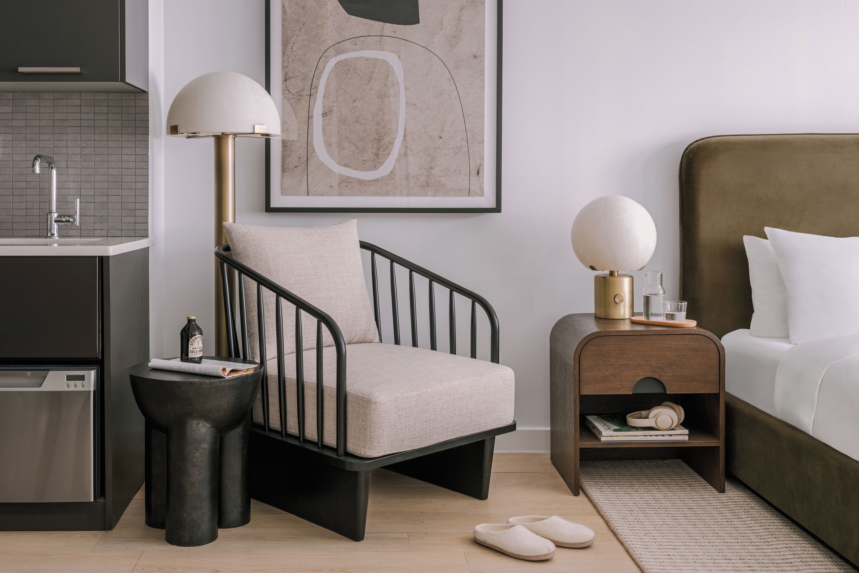 Un dormitorio moderno con cama, mesita de noche, lámpara, silla y una pequeña mesa con una botella y un libro.
