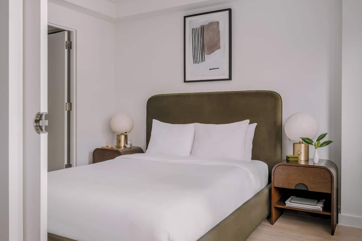Uma cama bem arrumada com cabeceira, duas luminárias laterais, uma obra de arte emoldurada na parede e uma mesa de cabeceira com uma planta.