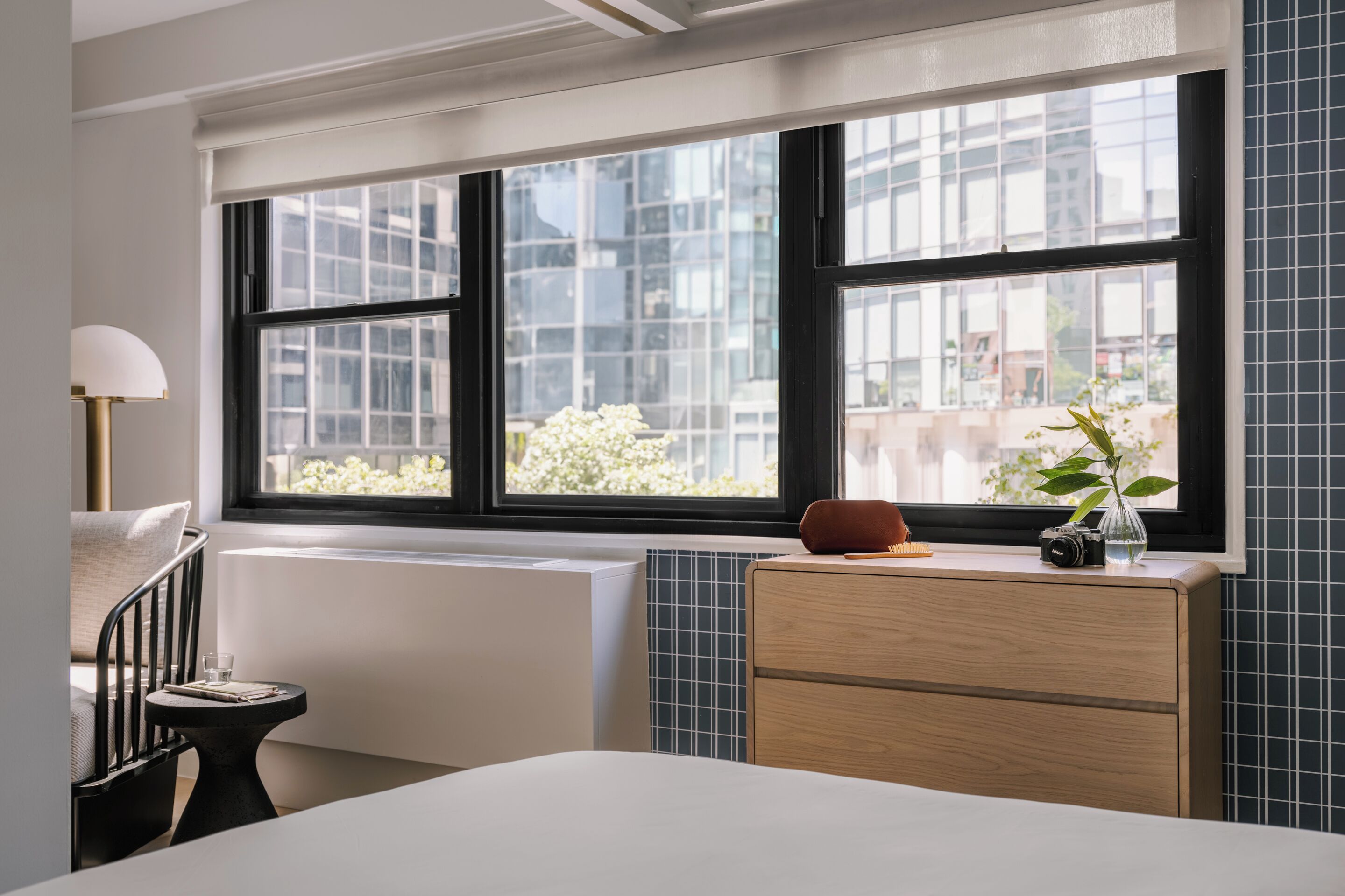 Une chambre d’hôtel avec une grande fenêtre, un radiateur, une commode en bois et une petite table avec une lampe.
