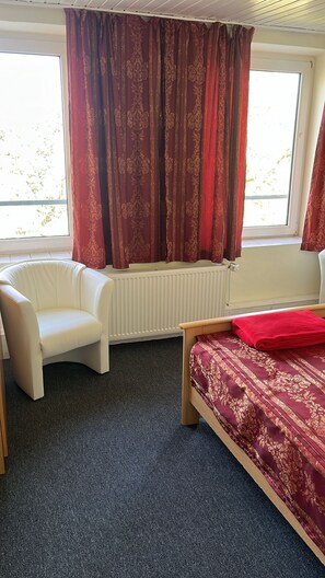 A room with a white chair, a bed with a red blanket, and a window with red curtains. - Vacation for the whole family (Malente)