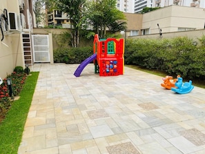 Área externa pavimentada com parquinho infantil, um escorrega pequeno e um cavalo de balanço azul.