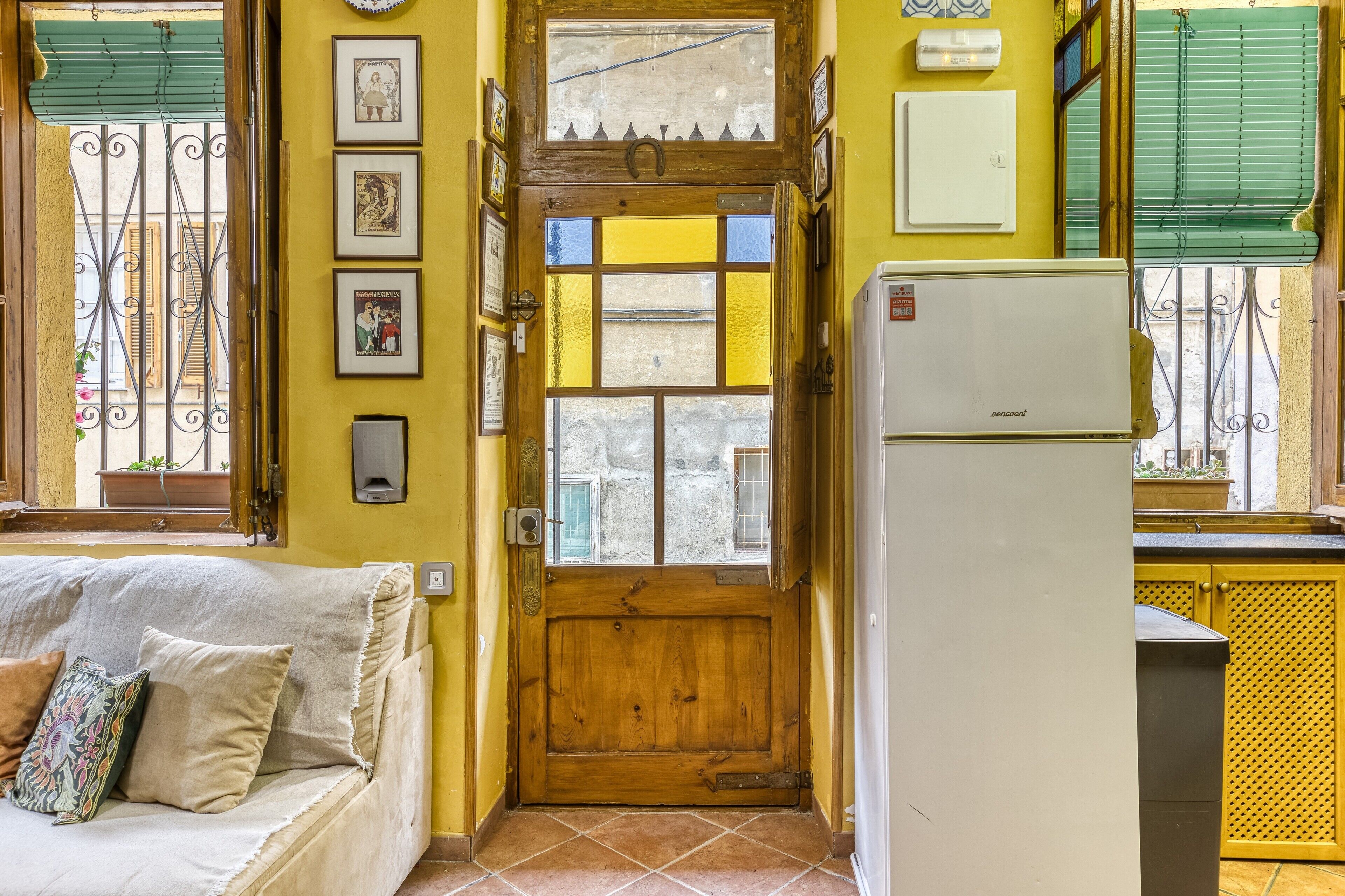 A room with a wooden door, a white refrigerator, a sofa with cushions, and a window with a view.