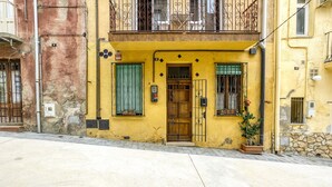 A yellow building with a wooden door, a balcony with metal railings, and a potted plant on the ground floor.