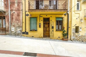 A yellow building with a wooden door, a balcony with metal railings, and a potted plant on the ground floor.