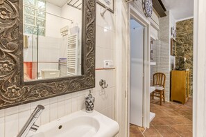 Een badkamer met een witte wastafel, een spiegel met een versierd omlijsting, een douche en een deur naar een kamer met een stoel en een kast.