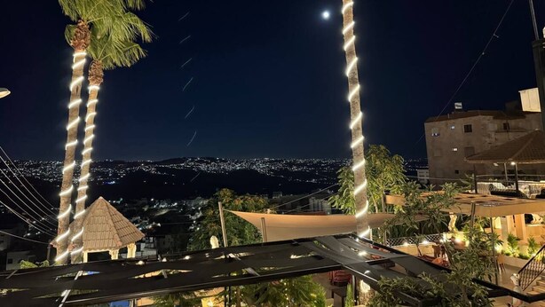 Deux palmiers illuminés par des guirlandes lumineuses, une petite construction dotée d’un toit de chaume et une vue sur un paysage urbain nocturne.