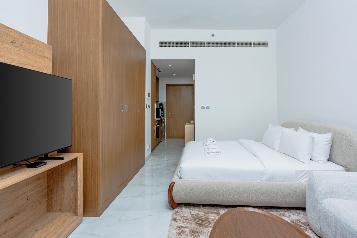 Habitación de hotel moderna con televisión de pantalla plana, armario de madera, cama con ropa de cama blanca y un pequeño rincón de descanso.