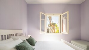 Un dormitorio con cama, almohadas, un banco y una ventana con contraventanas abiertas.