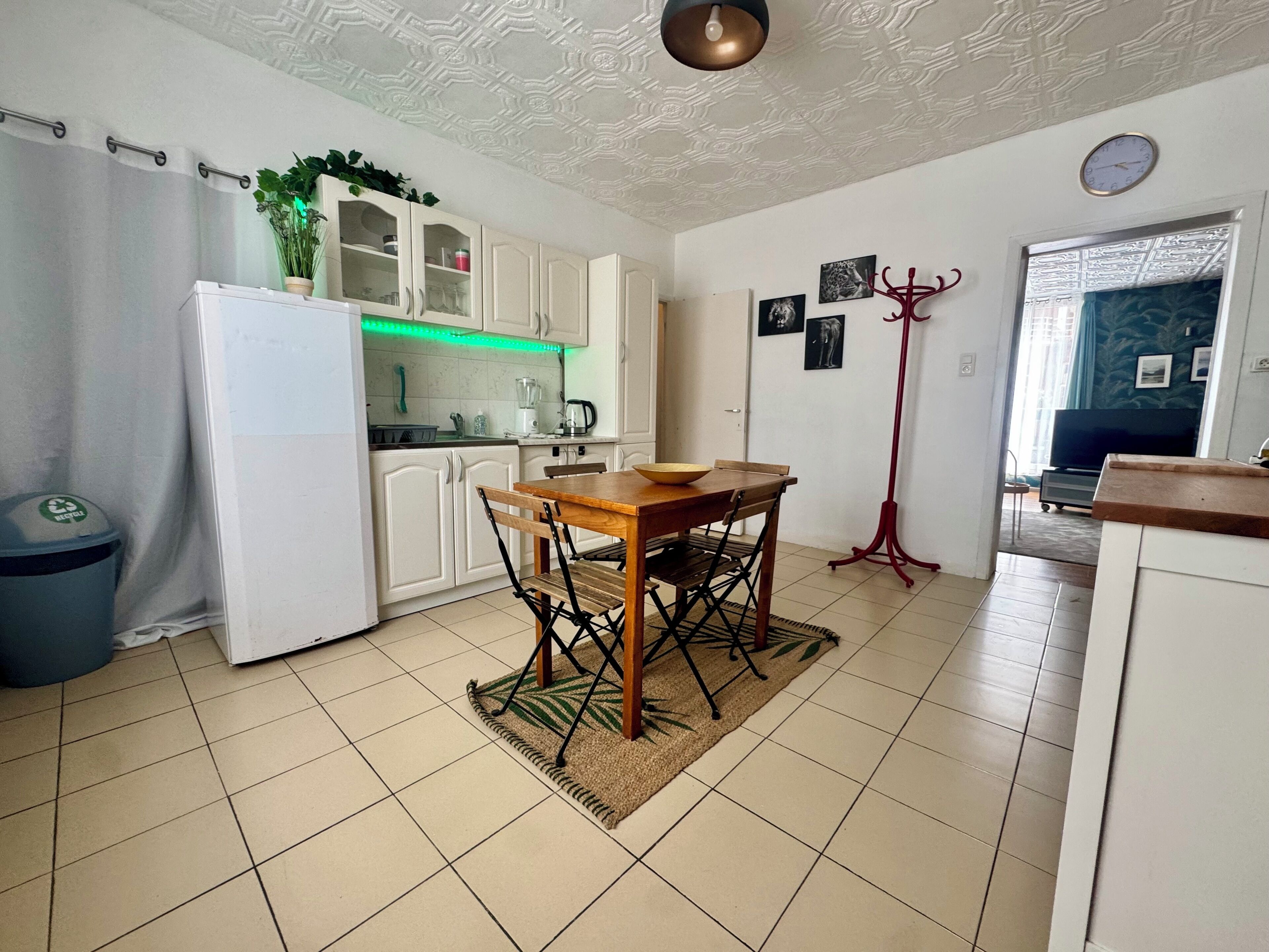 Une cuisine avec des meubles blancs, un réfrigérateur, une table à manger avec des chaises, une poubelle et un lampadaire.