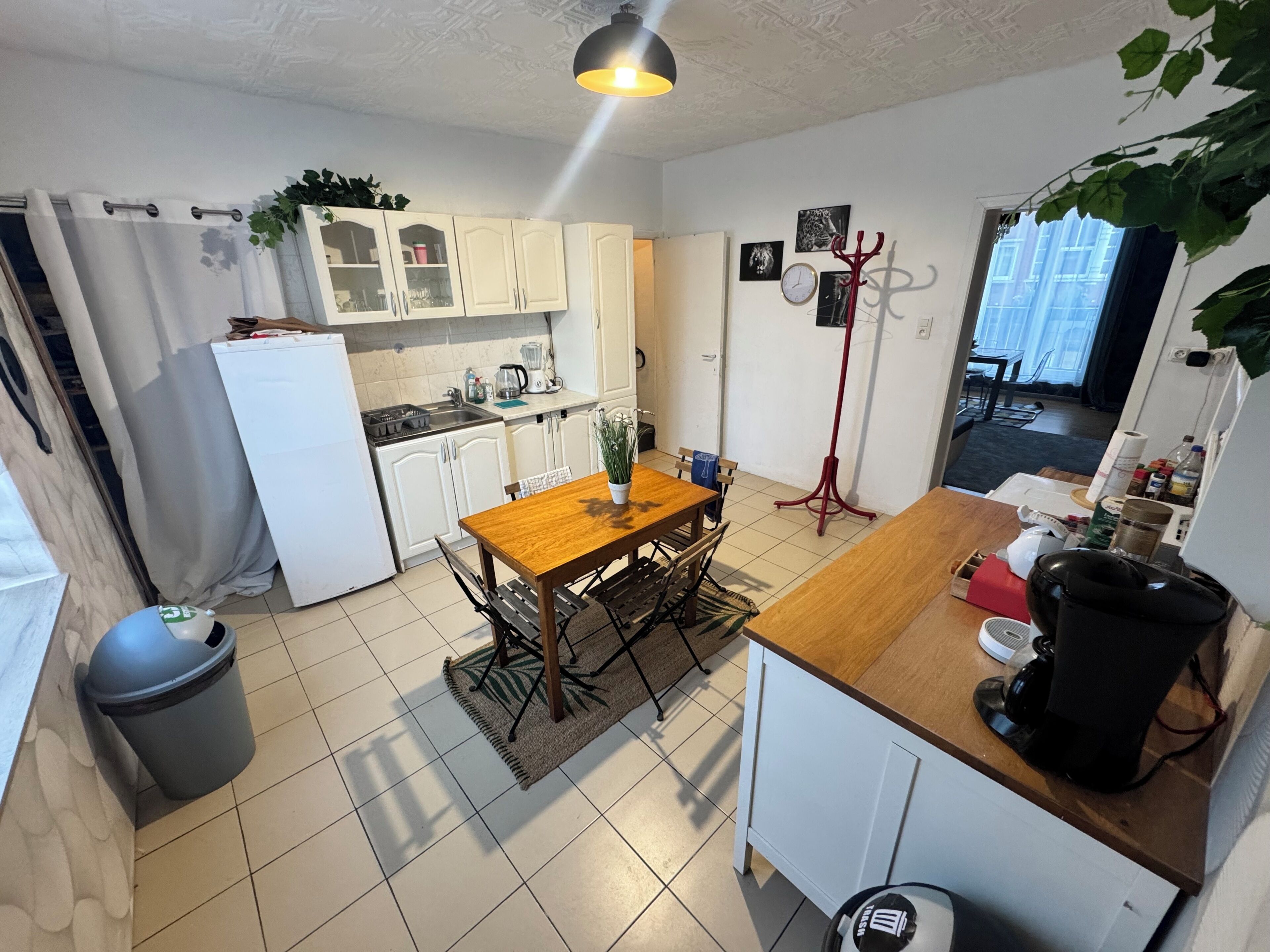 Een kleine keuken met witte kastjes, een houten eettafel, een koffieapparaat en een rode vloerlamp.