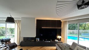 A modern living room with a flat-screen TV, a glass coffee table, a sofa, and a pool view.