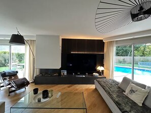 A modern living room with a flat-screen TV, a glass coffee table, a sofa, and a pool view.