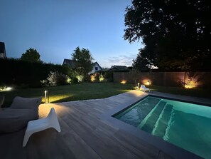 A backyard with a swimming pool, a wooden deck, and outdoor lighting.