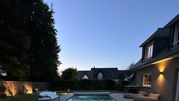 A rectangular swimming pool with a wooden deck in the foreground, a house with a dark roof and light exterior walls in the background, and a clear sky.