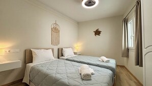 A room with two beds, a wall-mounted macrame decoration, a turtle wall art, a ceiling fan, and a window with curtains. - Kumquat Apartments (Corfu)