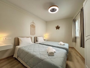 A room with two beds, a wall-mounted macrame decoration, a turtle wall art, a ceiling fan, and a window with curtains. - Kumquat Apartments (Corfu)