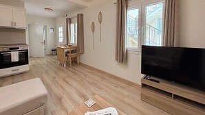 A modern living room with a flat-screen TV, wooden flooring, a dining area with a table and chairs, and a kitchen with cabinets and an oven. - Kumquat Apartments (Corfu)