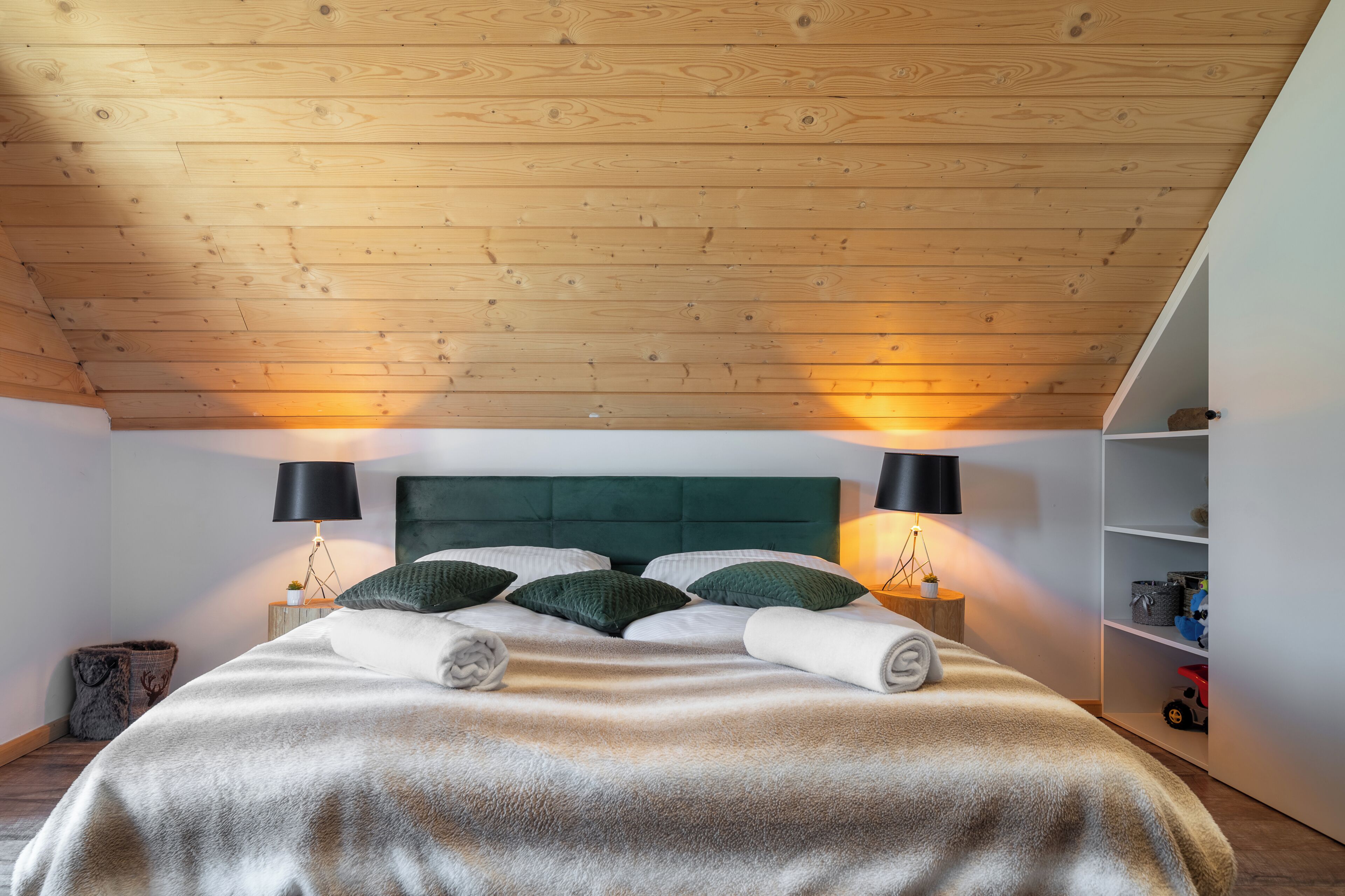Ein Schlafzimmer mit Holzboden, einem grünen, gepolsterten Kopfteil, zwei Nachttischlampen, einem Bett mit weißer Decke und einem weißen Kleiderschrank mit Regalen.