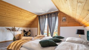Un dormitorio con techo inclinado, una cama con ropa de cama blanca y almohadas verdes, una mesita de noche con una lámpara y una silla.