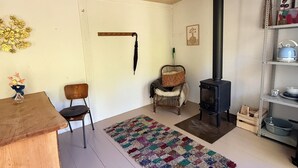 Un salón acogedor con una mesa de madera, una silla, un tapete colorido, una butaca de mimbre, una estufa negra y una estantería metálica con objetos.
