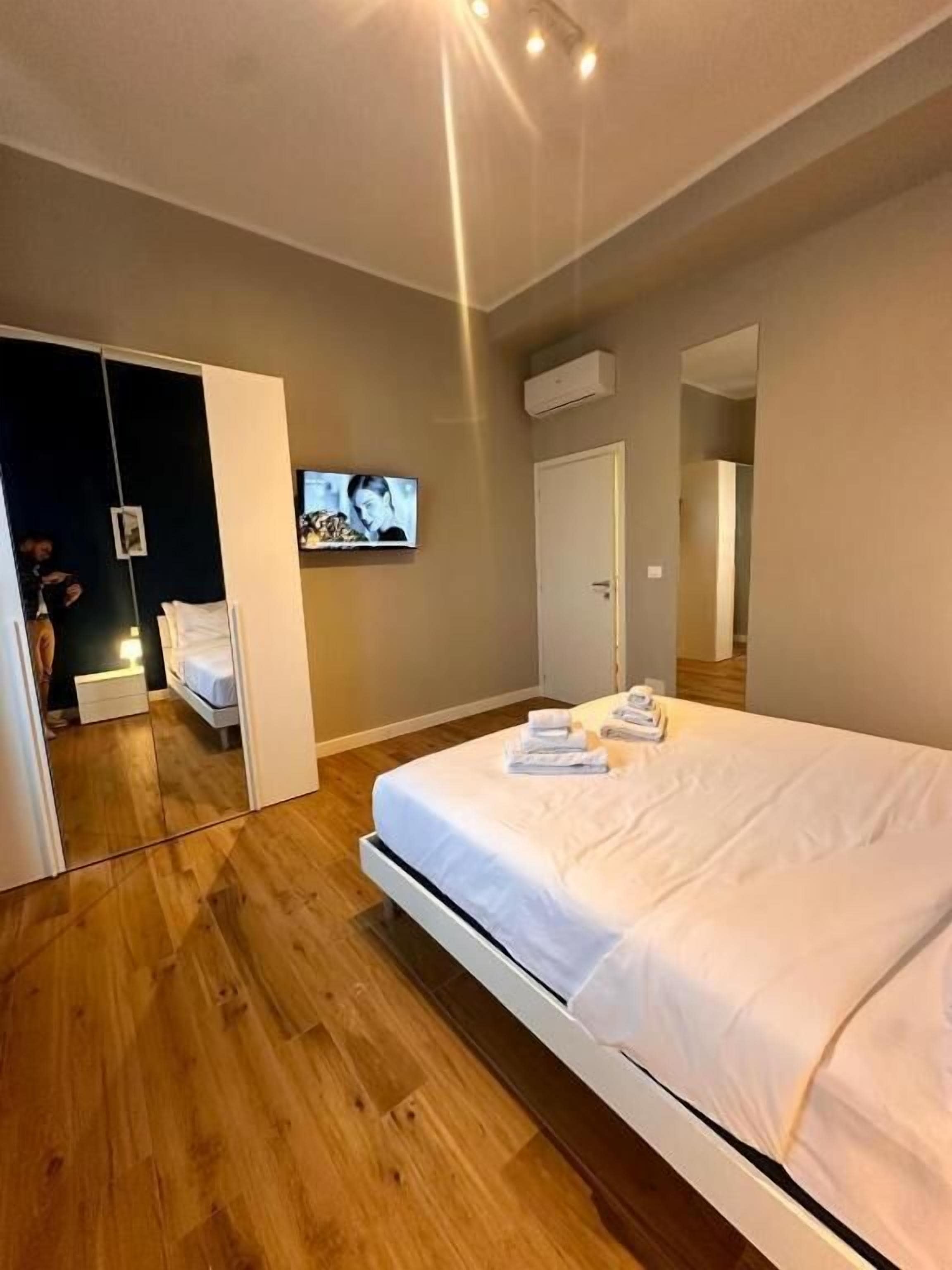A modern hotel room with a large bed, wooden flooring, a flat-screen TV mounted on the wall, and a wardrobe with a mirror.