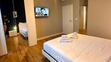 A modern hotel room with a large bed, wooden flooring, a flat-screen TV mounted on the wall, and a wardrobe with a mirror.