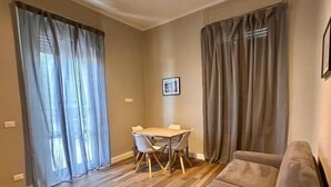 A living room with a wooden floor, a sofa, a dining table with chairs, and curtains on the windows. - HH Tortona (Tortona)