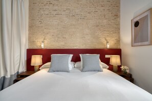 Een hotelkamer met een bakstenen muur, een rode hoofdbord, twee nachtlampjes, een bed met witte lakens en een ingelijst schilderij aan de wand.