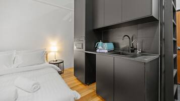 A compact room with a bed, bedside lamp, kitchenette, and a small table.