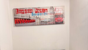 Una cama con una colcha estampada, dos almohadas y una toalla doblada en forma de cisne. En la pared sobre la cama hay una ilustración de un autobús de dos pisos rojo.
