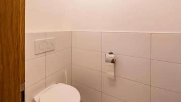 A small, clean bathroom with a toilet, a paper towel dispenser, and a wall-mounted flush button.