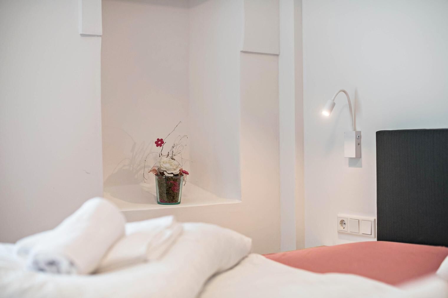 A hotel room with a bed, a bedside table with a vase of flowers, a wall-mounted light, and a power outlet.