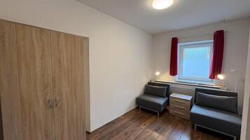 A room with a wooden wardrobe, two grey sofas, a small table, and a window with red curtains.