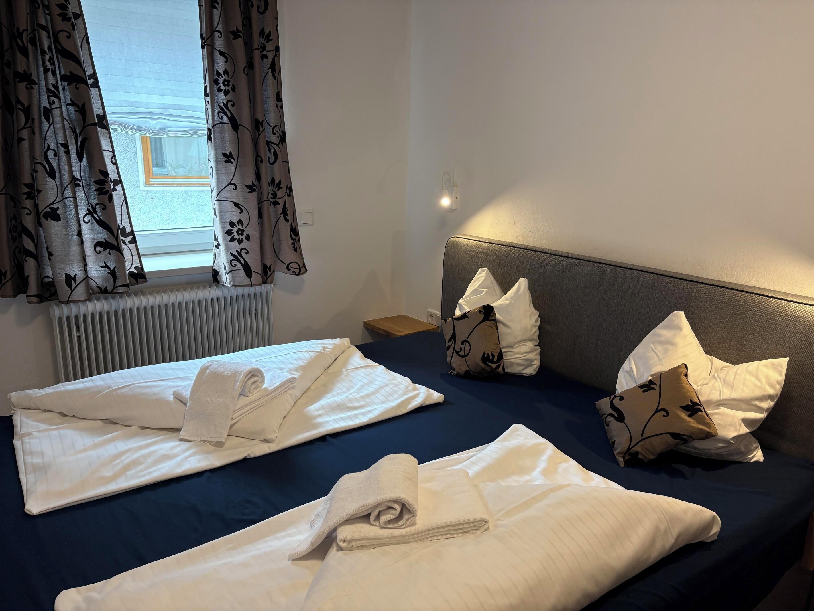 A hotel room with two beds, each with neatly folded towels and pillows, a radiator, a window with curtains, and a bedside table.