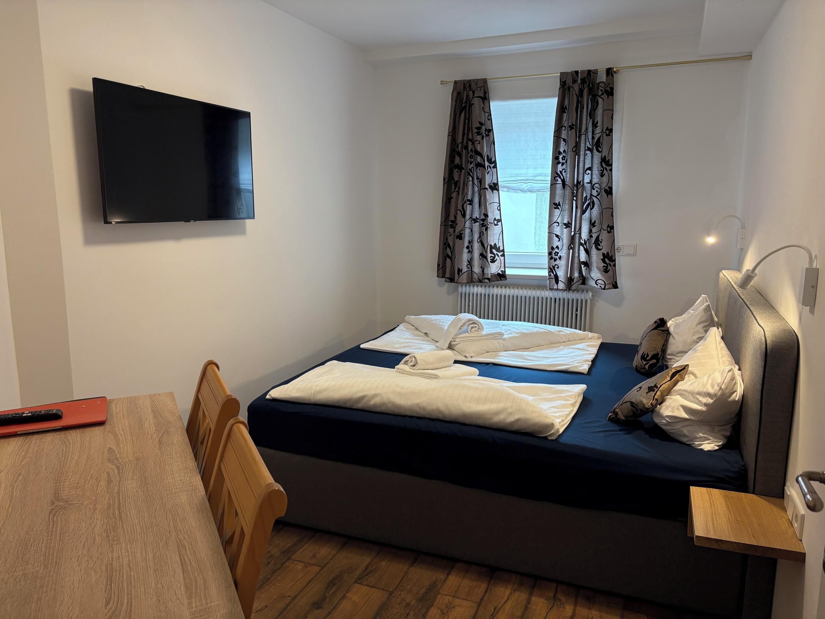 Une petite chambre d’hôtel comprenant un lit, un bureau, une chaise, un téléviseur fixé au mur et une fenêtre avec des rideaux.