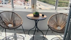 A balcony with two wicker chairs, a small table with a plant, and a view of a road and buildings. - Bright one bedroom apartment in La Mata, Avenida Suiza. With pool. (La Mata)