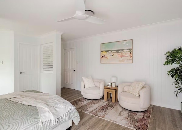 A bedroom with a bed, two armchairs, a small table, a ceiling fan, a painting, and a potted plant.