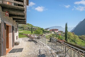 Un balcón con suelo de piedra, mobiliario exterior, barandilla de metal y vistas a un paisaje montañoso.