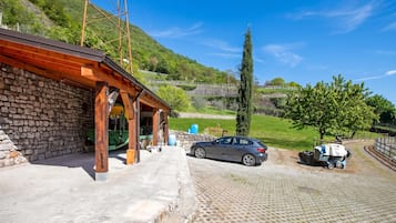 A stone building with a wooden roof, a parked car, and a paved area with a tree and a fence.