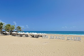 Une plage avec des chaises longues et des parasols, une pancarte indiquant « THE BEACH CLUB », et des palmiers.
