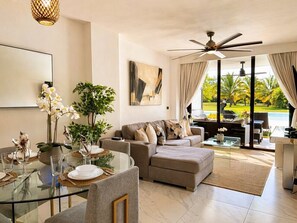 Un salon moderne avec une table à manger en verre, un canapé, un ventilateur de plafond et une vue sur un jardin grâce à de grandes fenêtres.
