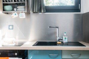 Une cuisine équipée d’un évier en acier inoxydable, d’une fenêtre située au-dessus de l’évier et d’un égouttoir contenant des tasses et des bols.