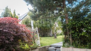 Eine Steintreppe führt zu einem Garten mit einem weißen Tor, umgeben von viel Grün und einem Gebäude im Hintergrund.