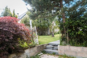A stone staircase leading to a garden with a white gate, surrounded by greenery and a building in the background.