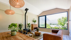 A modern living room with a wooden dining table, leather sofa, and large windows offering a mountain view. - Hemelberg Guesthouse (Stellenbosch)