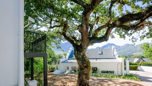A tree-lined pathway leading to a building with a metal roof, surrounded by mountains. - Hemelberg Guesthouse (Stellenbosch)