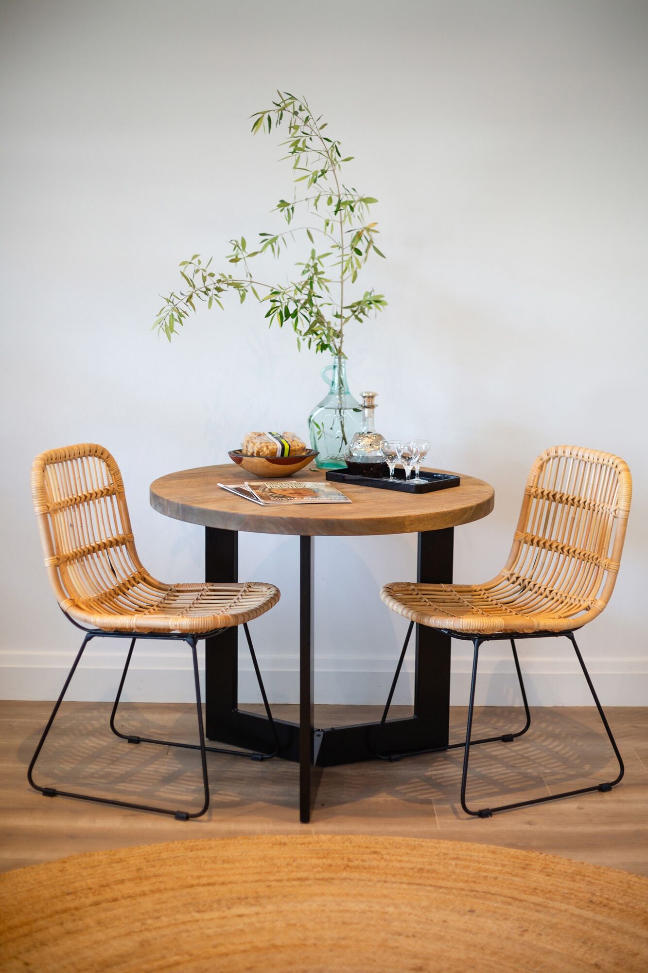 A round table with two wicker chairs, a vase with a plant, a bowl of bread, and a tray with glasses.
