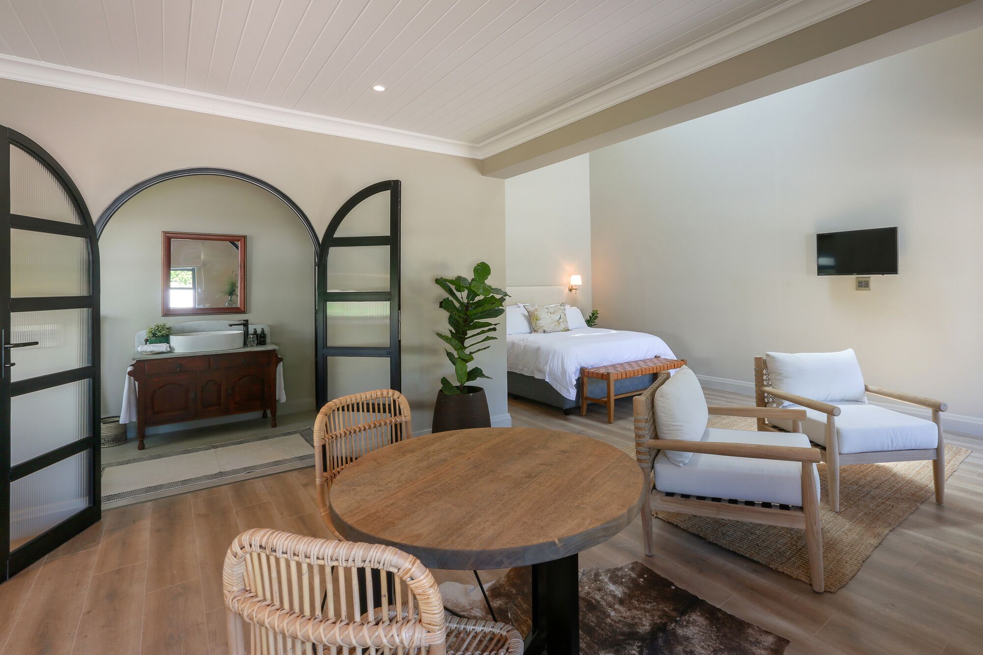 Ein Zimmer mit einem Bett, zwei Korbstühlen, einem runden Holztisch, einem an der Wand befestigten Fernseher und einem Badezimmer mit Spiegel und Waschbecken.