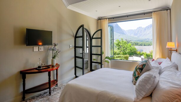 A hotel room with a bed, a TV mounted on the wall, a small table with a vase and a tray, and a window with a mountain view. - Hemelberg Guesthouse (Stellenbosch)