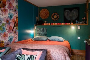 Une chambre avec un mur de couleur turquoise, un papier peint à motifs floraux, un lit recouvert d’une courtepointe orange, deux oreillers, une étagère avec des objets décoratifs et un miroir fixé au mur.